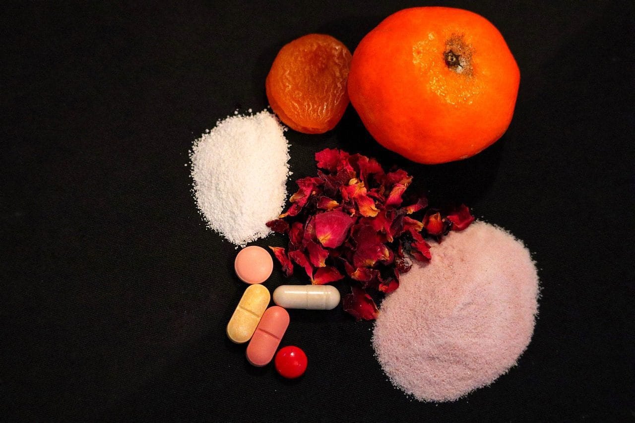A variety of health supplements including tablets, capsules, and pink and white powders arranged with a fresh orange and dried fruit