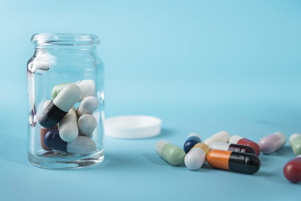 Assorted colorful health supplement capsules scattered on a blue surface next to a small clear glass apothecary jar