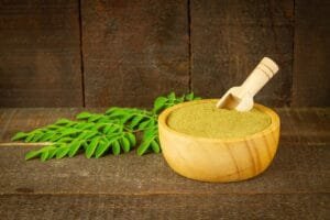 Green herbal moringa powder in a wooden bowl with a small scoop, placed next to a fresh green leaf branch on a rustic wooden table