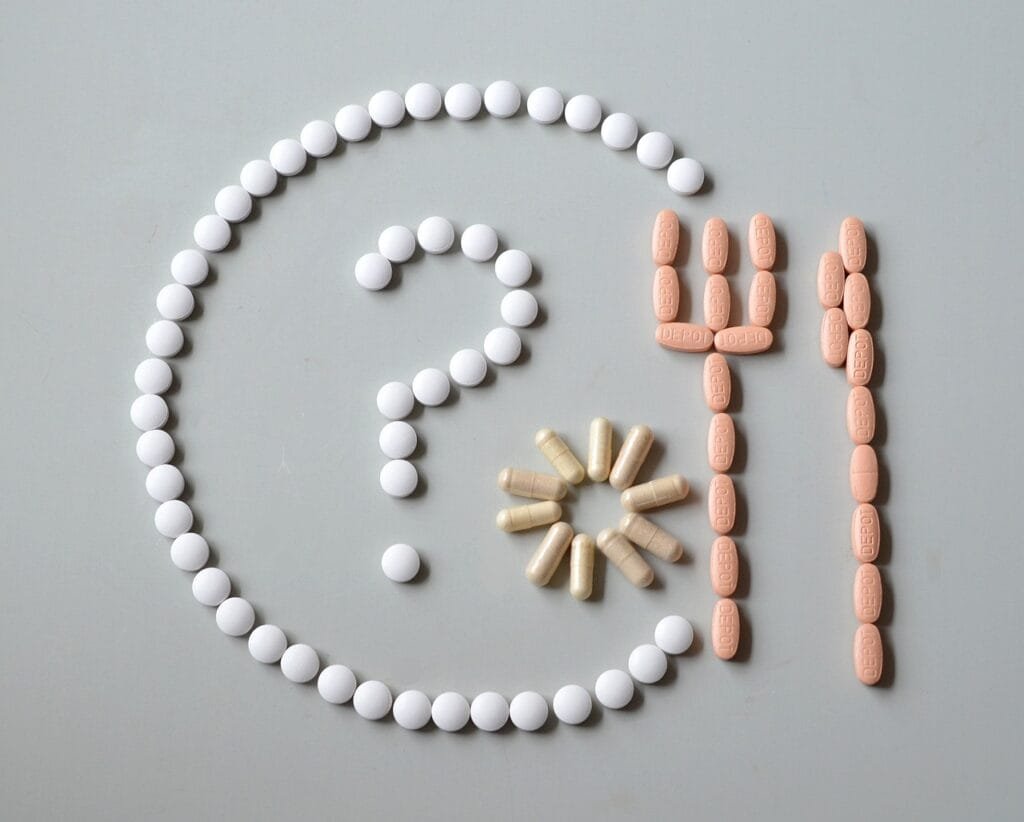 Various colorful supplement tablets and capsules arranged on a gray surface to form a question mark, fork, and knife