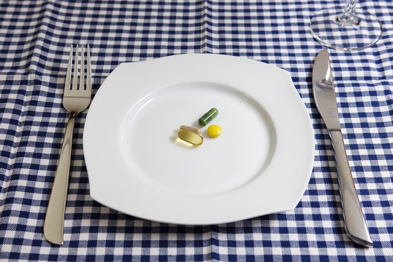 A small assortment of health supplement capsules and tablets centered on a white plate with a fork and knife on a blue checkered tablecloth