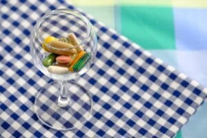 A variety of colorful health supplement capsules and tablets inside a clear wine glass on a blue checkered tablecloth