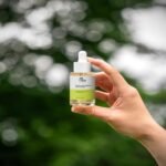 A close-up of a person's hand holding a clear 30ml bottle of Vita Elixia Bioactive Prebiotics Jelly Serum against a soft, blurred background of lush green trees