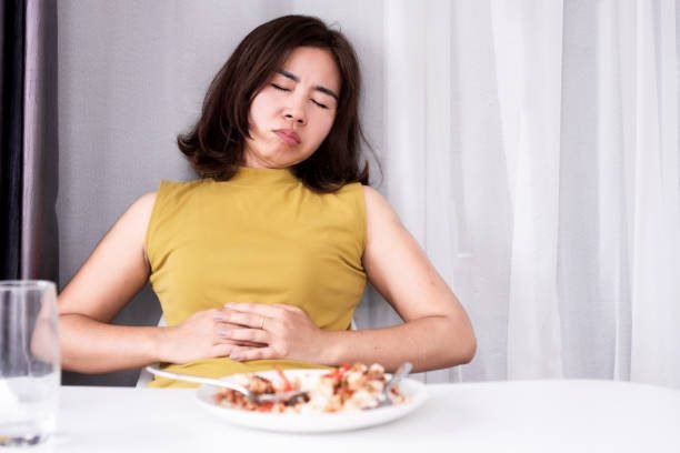 A woman in a yellow shirt holding her stomach in discomfort while eating, illustrating digestive health and gut wellness tips