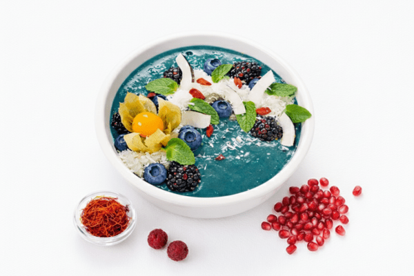 A vibrant blue spirulina smoothie bowl topped with blueberries, blackberries, coconut flakes, mint, and a physalis fruit, served with saffron and pomegranate seeds