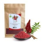 A 200g brown paper pouch of Blumental Organic Sumac next to a small bowl of deep-red spice powder and a fresh staghorn sumac branch.