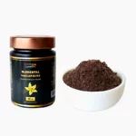 A premium 15g black glass jar of Blumental Vanillepulver with a copper-colored lid, displayed alongside a small white bowl of 100% pure ground vanilla