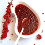A white, kidney-shaped bowl filled with vibrant red organic barberry jam, shown with a spoon and a fresh branch of bright red barberries on the side