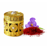 An ornate, gold-embossed cylindrical gift box decorated with pearls, displayed next to a pile of deep-red saffron threads and a purple saffron crocus flower.