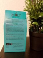 Teal packaging for Moly "Defence" herbal tea, showing the back label with descriptions in German and English, placed next to a small green potted plant.