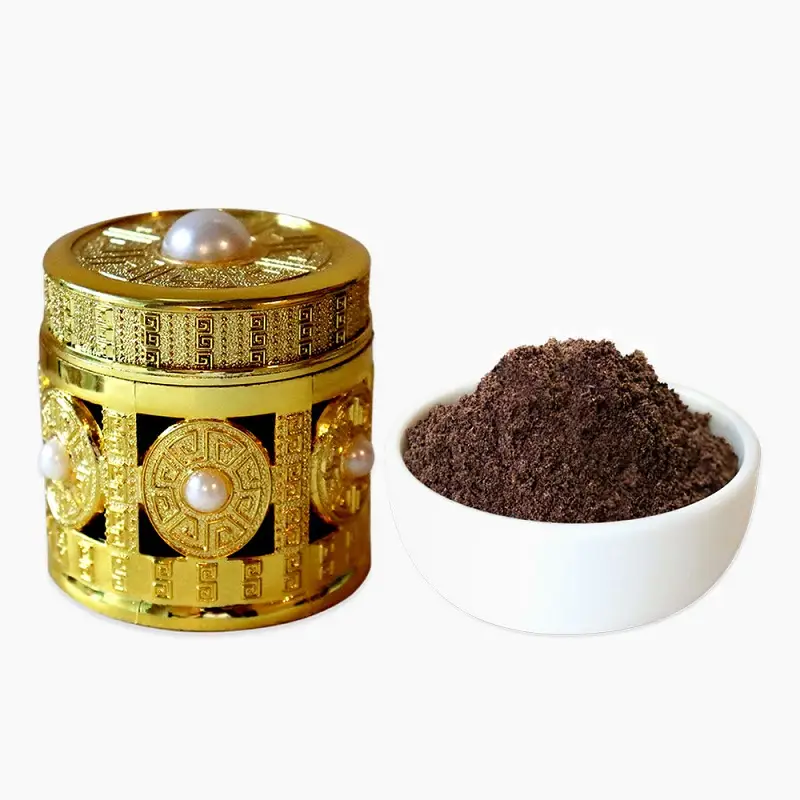 An ornate, gold-embossed cylindrical gift box decorated with pearls, displayed next to a small white bowl filled with finely ground, aromatic black vanilla powder.