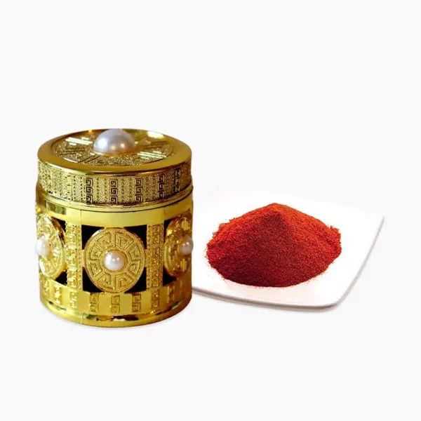 An ornate, gold-embossed cylindrical gift box decorated with pearls, displayed next to a small white dish containing finely ground, vibrant red saffron powder.